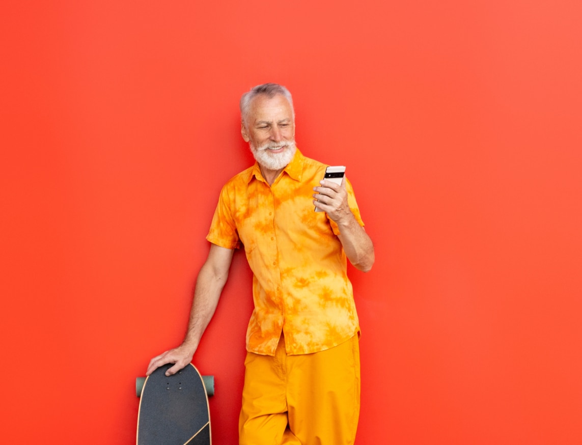 Smiling handsome senior man holding skateboard and mobile phone reading text message, shopping online isolated on red background. Bearded hipster using smartphone, communication online  on the street