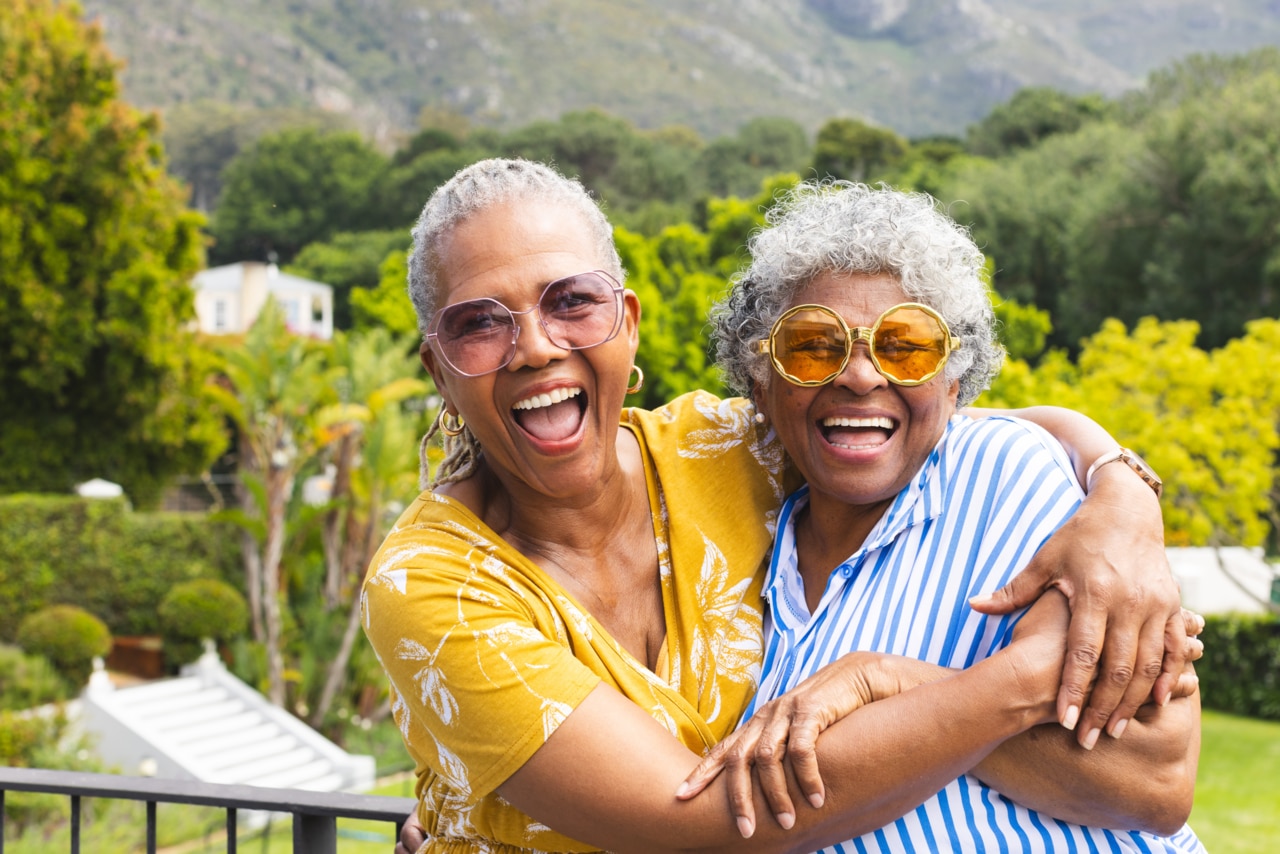 Two senior women share joyful hug outside. One wears yellow glasses