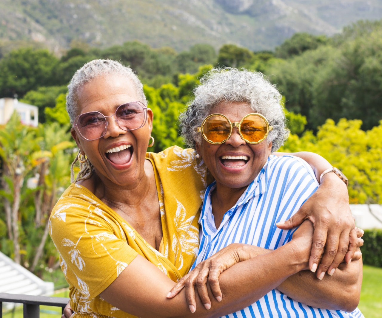 Two senior women share joyful hug outside. One wears yellow glasses