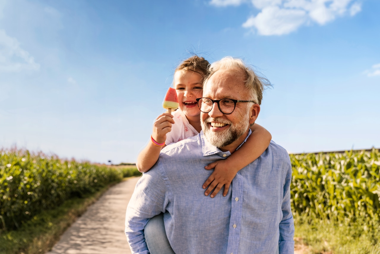 Grand-père souriant portant sa petite-fille sur son dos sur un chemin ensoleillé dans un champ de maïs, alors qu’elle tient une glace à la pastèque