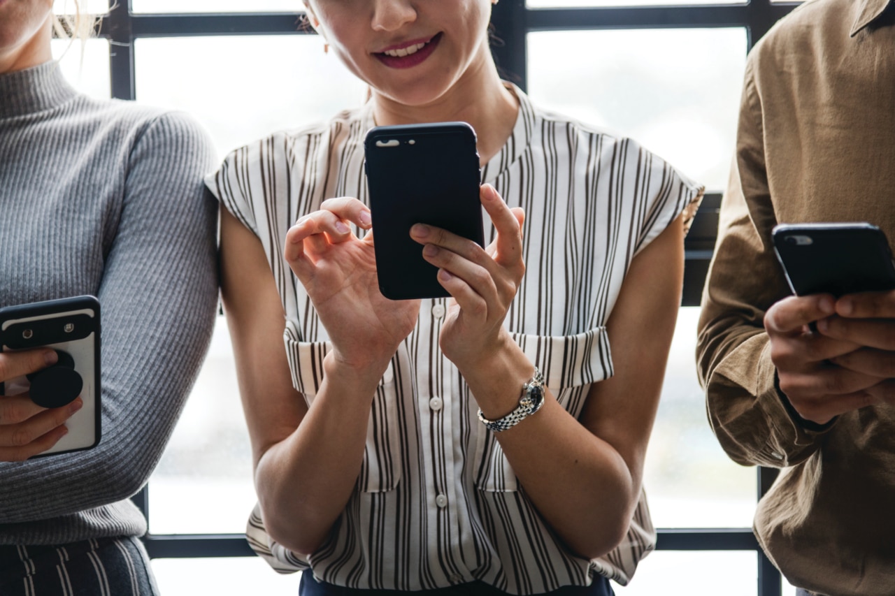 Un groupe de trois personnes debout, proches les unes des autres, chacune tenant un smartphone. La personne au centre se concentre sur son appareil, avec un léger sourire, tandis que les autres sont également occupées avec leur téléphone. Elles sont habillées de façon décontractée, avec des motifs et des couleurs variés