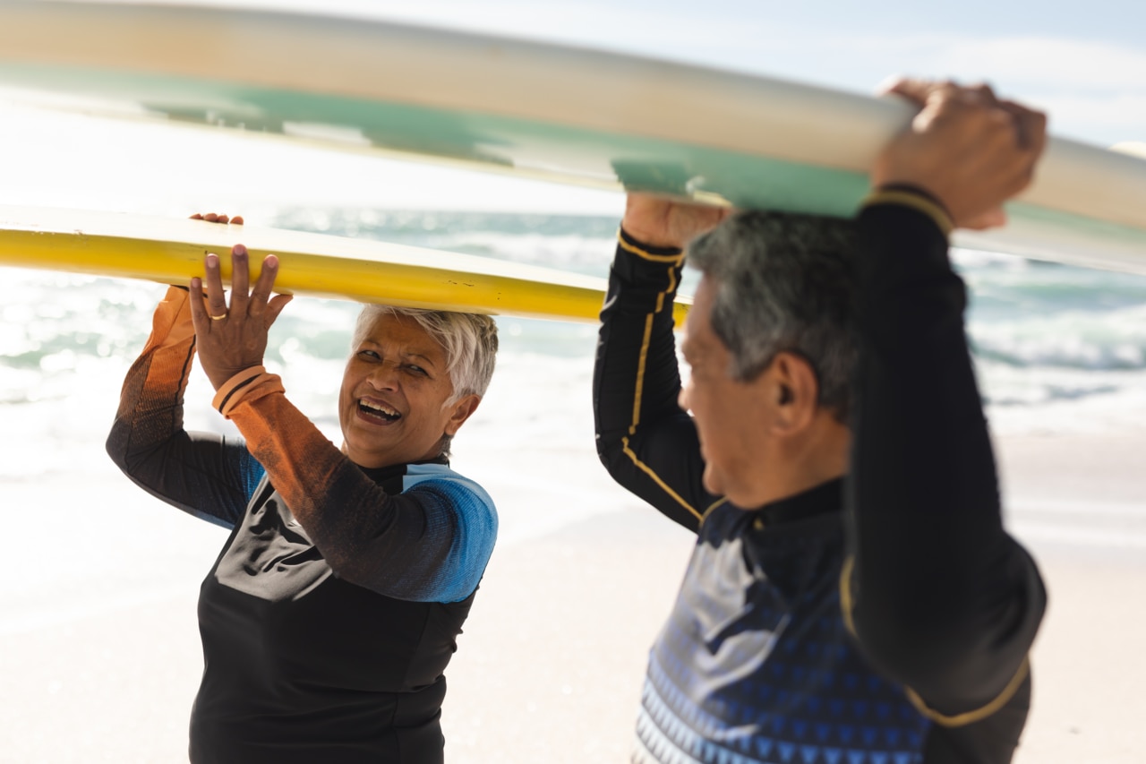 Mulher sênior alegre, birracial, carregando uma prancha de surfe sobre a cabeça enquanto olha para um homem que faz o mesmo. Esporte aquático e estilo de vida ativo.