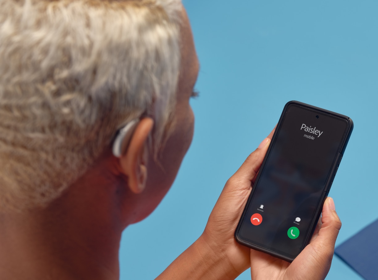 Uma mulher negra, de cabelos brancos, usando aparelho auditivo e segurando um smartphone que exibe uma chamada recebida, sugerindo a conexão entre o aparelho auditivo e um aplicativo remoto.