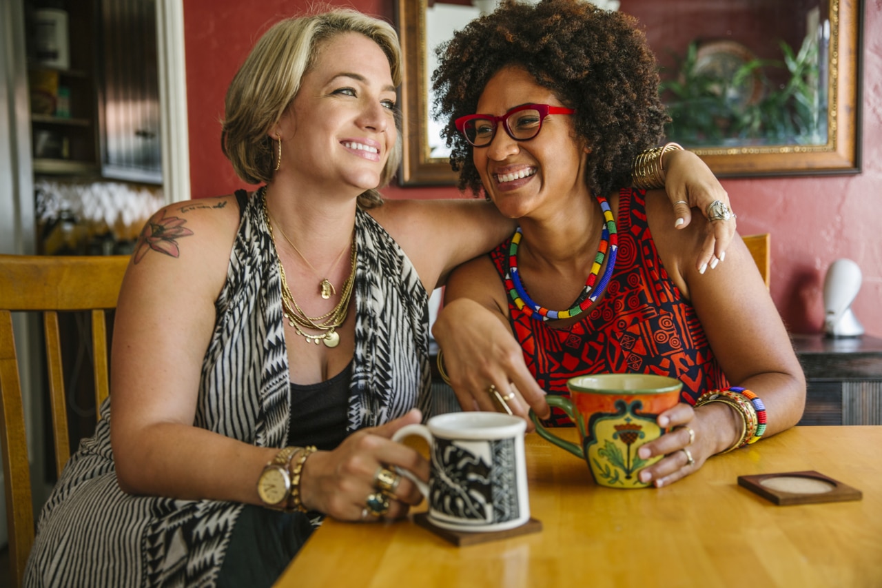 Duas mulheres estão sentadas juntas a uma mesa de madeira, sorrindo e aproveitando a companhia uma da outra. Uma delas está usando uma blusa estampada com colares em camadas, enquanto a outra está vestida com uma roupa colorida e vibrante e adornada com joias. Cada uma delas está segurando uma caneca estilizada.