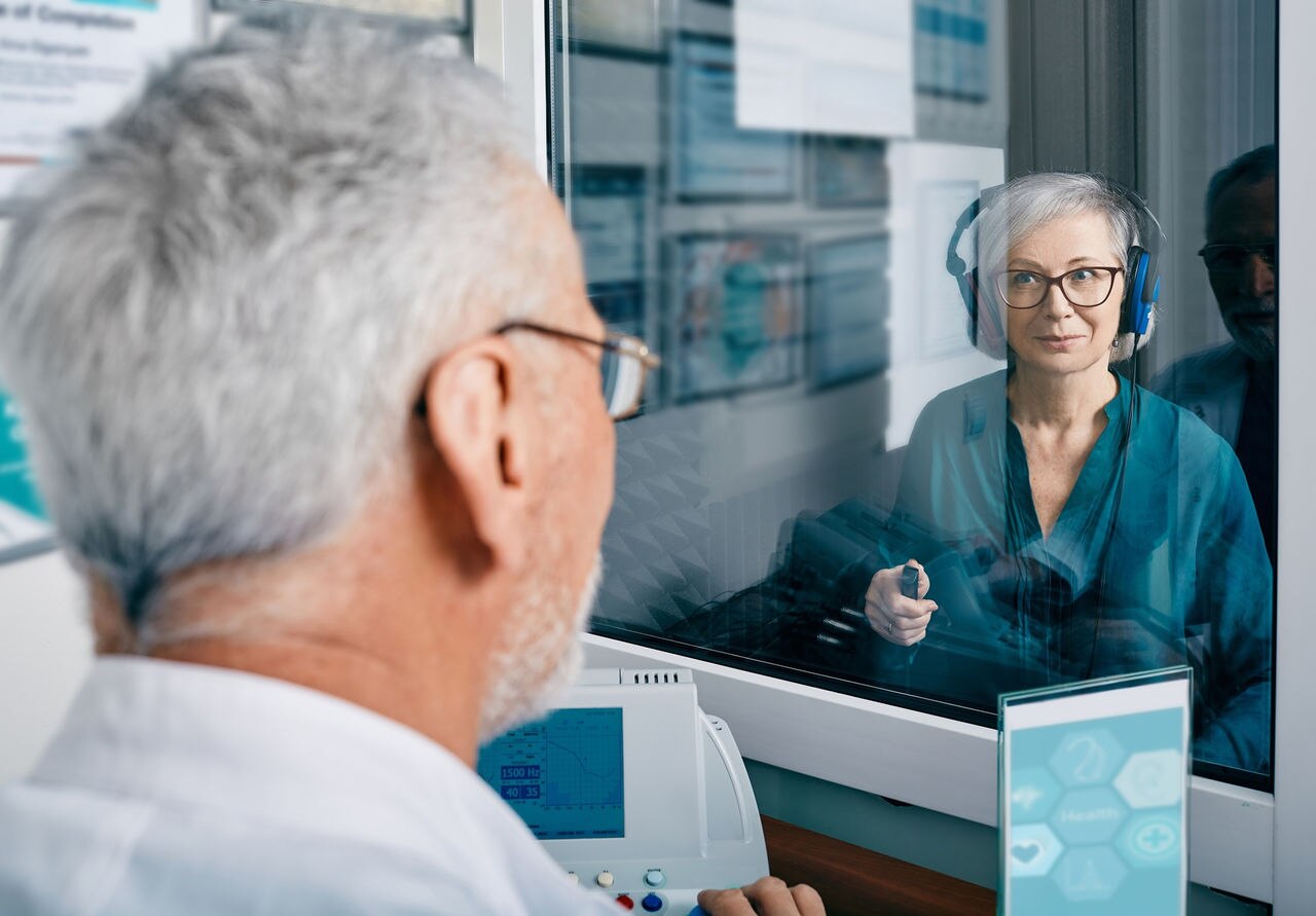 A healthcare professional sitting at a desk, facing a patient who is wearing headphones and sitting behind a glass partition, with various medical charts and equipment visible in the background