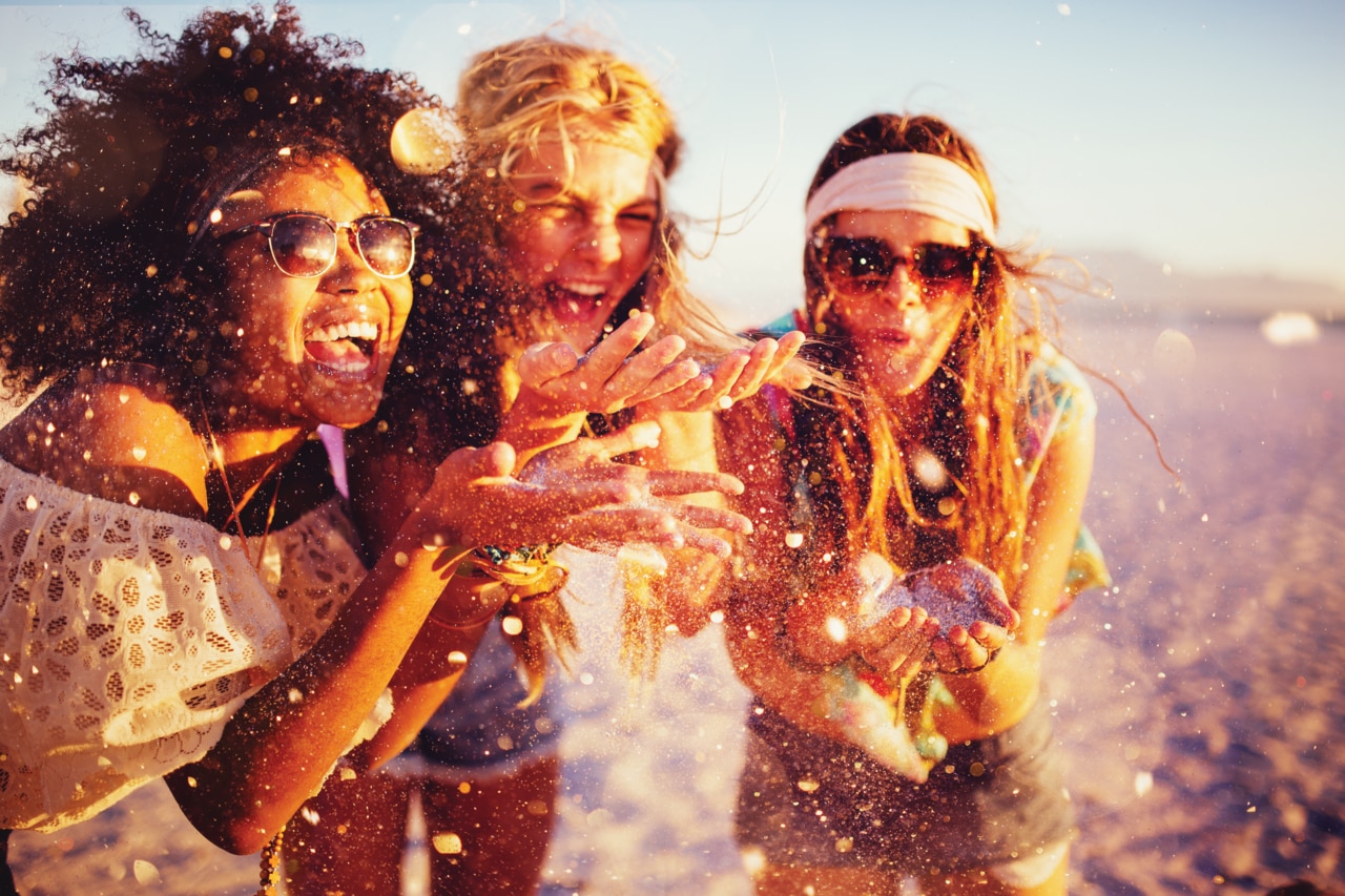 Mixed race group of girls blowing colourful confetti from their hands happily on a beach at sunset