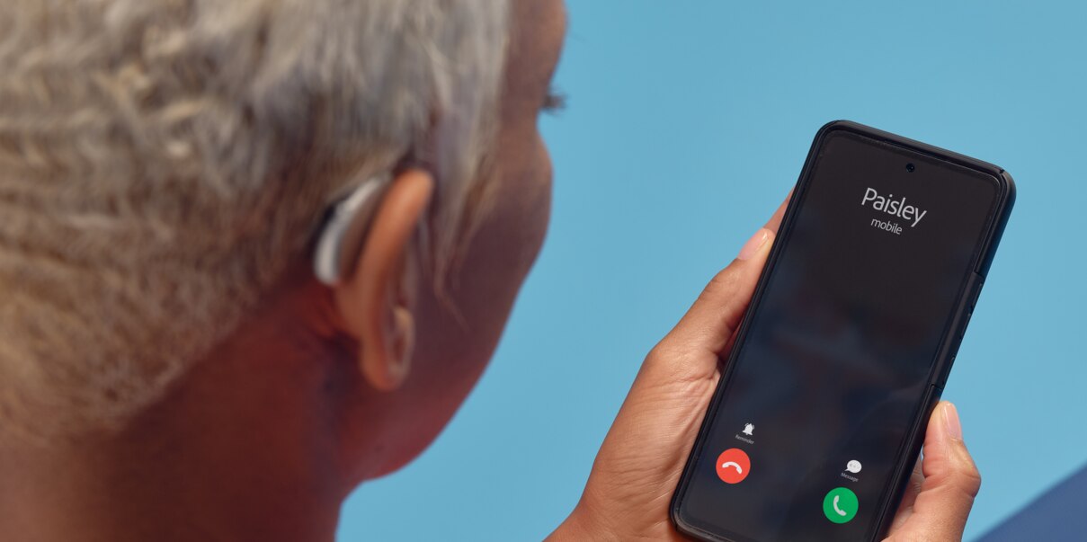 A female, white-haired African hearing aid wearer holding a smartphone that displays an incoming call., suggesting a connection between hearing aids and remote app.