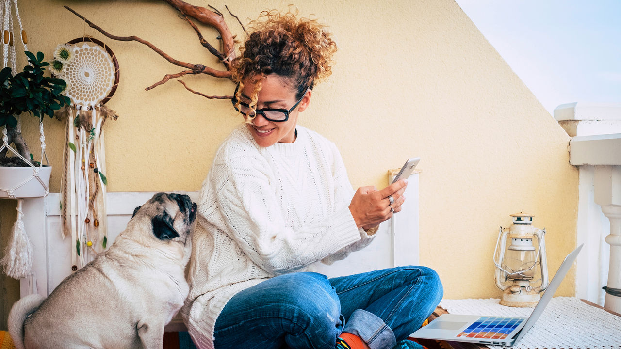 Une femme et son adorable carlin sont assis chez eux pour travailler avec des appareils technologiques comme un téléphone et un ordinateur portable.