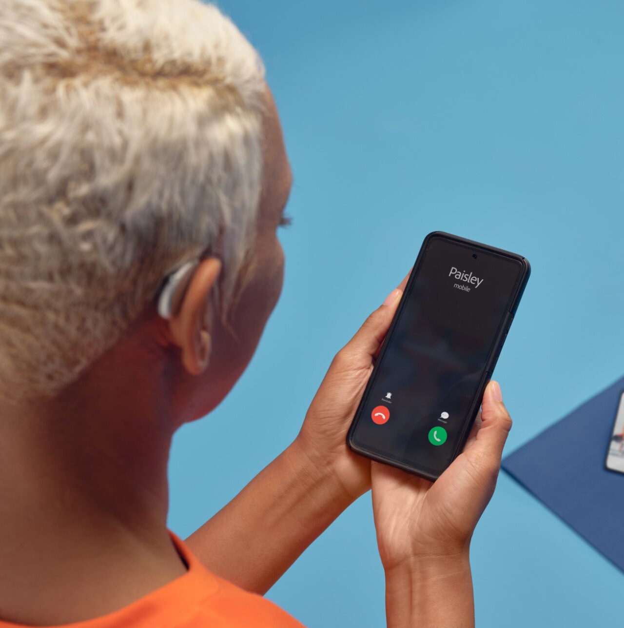 Une femme aux cheveux blancs, portant un instrument auditif et tenant un téléphone intelligent qui affiche un appel entrant, suggérant une connexion entre les instruments auditifs et une application à distance.