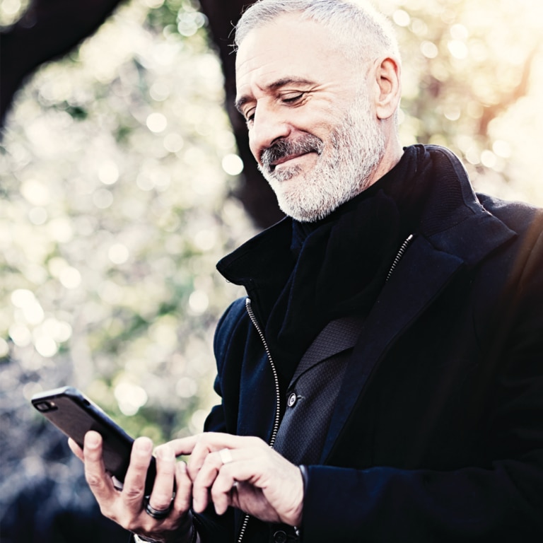 A person is standing outdoors, holding a smartphone in their hands while wearing a dark jacket. 
