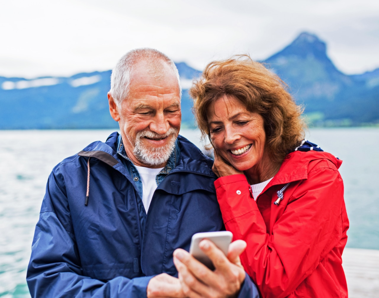  a couple standing close together by a body of water, likely a lake, with mountains in the background.