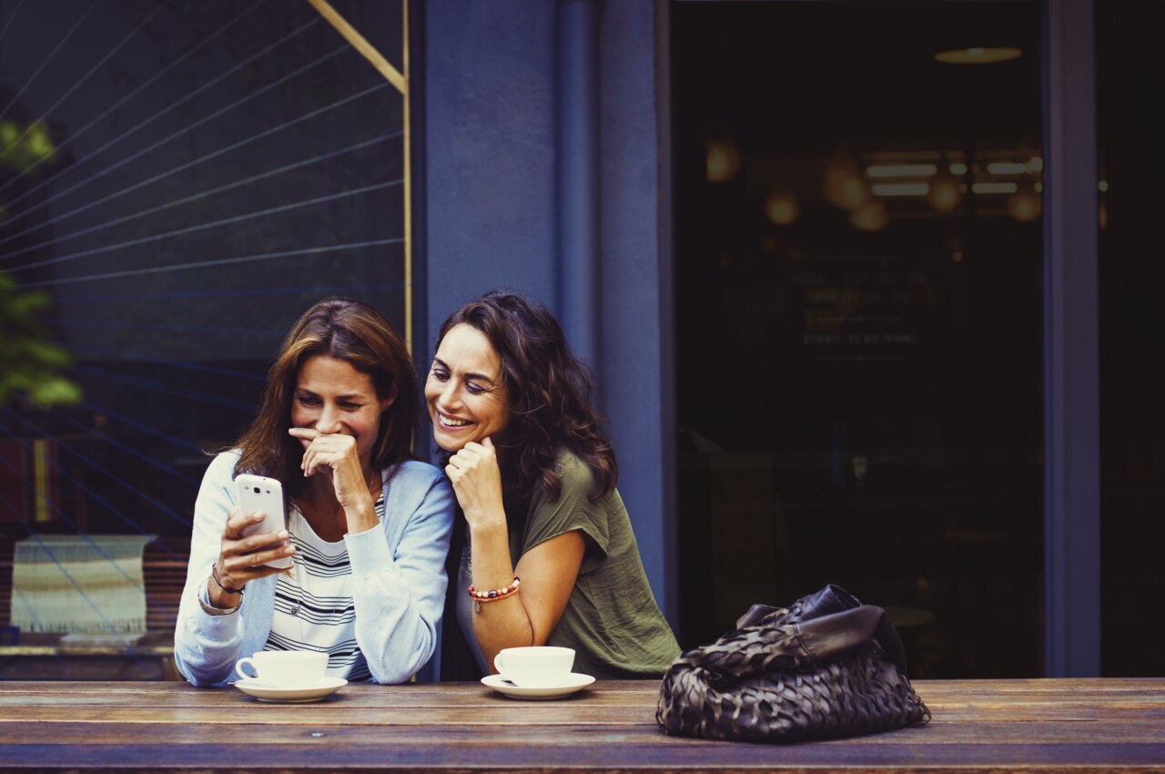 To venner sidder ved et træbord i en café og deler et øjeblik sammen. Den ene kigger på en smartphone, mens den anden læner sig tæt ind, begge smiler og nyder deres tid. To kopper kaffe er placeret på bordet, hvilket bidrager til den afslappede atmosfære.