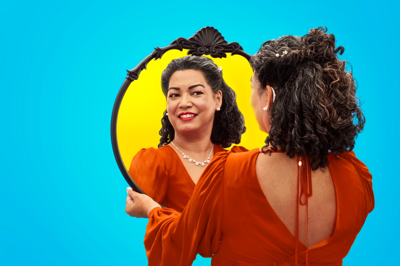 Una persona está de pie frente a un fondo azul vibrante, sosteniendo un espejo decorativo que refleja su imagen. Lleva un vestido elegante de color naranja con un diseño único en la espalda. El marco del espejo es decorativo, lo que añade un toque de elegancia a la escena. 