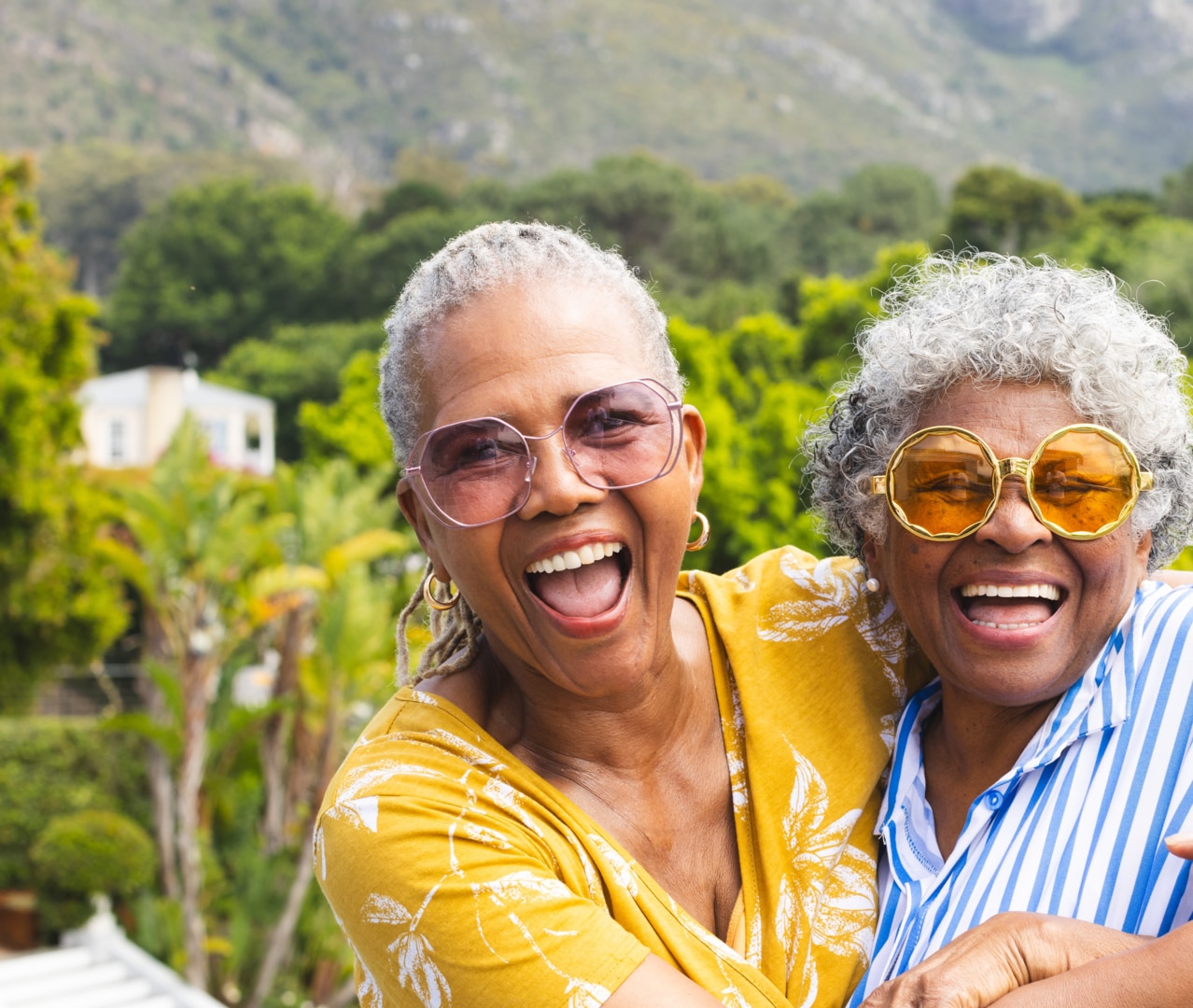 Two senior women share joyful hug outside. One wears yellow glasses