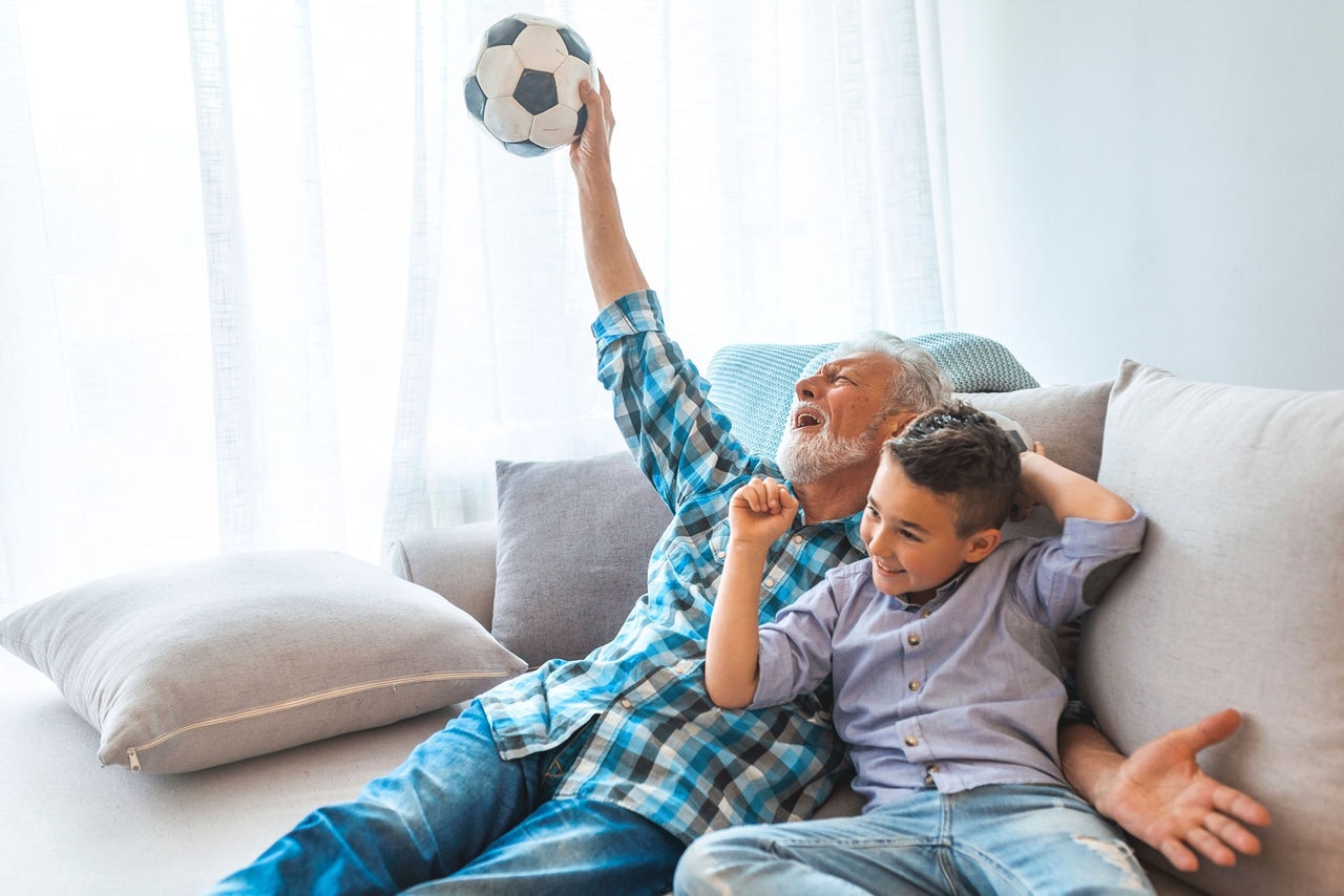 Excited grandchild and senior man watching football match. Overjoyed grandfather and kid watching football match together, cheering for a football game and holding a football ball ; Shutterstock ID 1418482811; purchase_order: cost center; job: 51700; client: Krista Koktan; other: Unitron