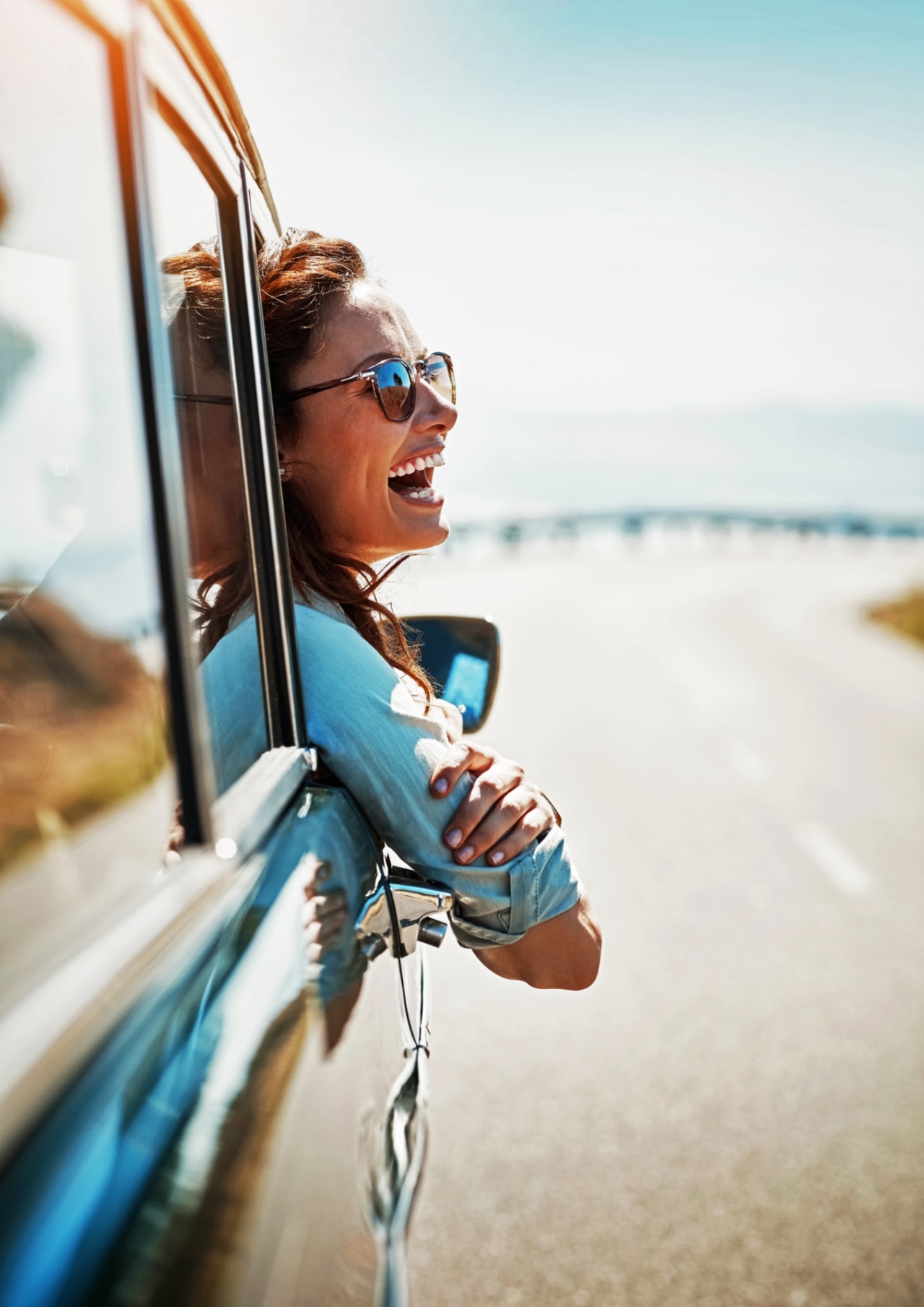 A woman leaning out of a car window, enjoying the view of a winding road ahead. The sunlight casts a warm glow, highlighting the person's relaxed posture and casual attire. The landscape in the background suggests a scenic drive, with open roads and distant hills.