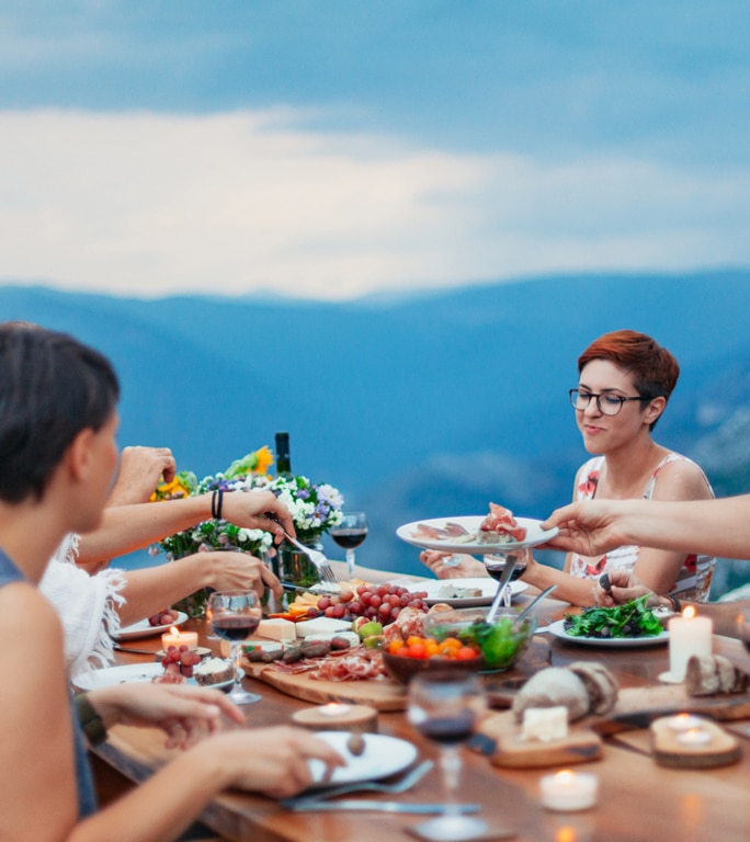 group of people is gathered around a beautifully set table outdoors, enjoying a meal with a stunning mountain view in the background. The table is adorned with various dishes, including salads, fruits, and meats, along with glasses of wine.