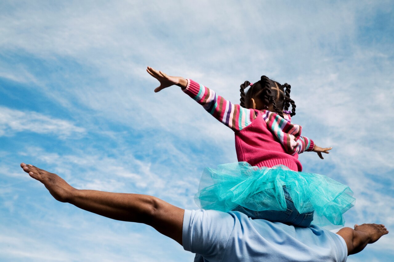 A young girl, wearing a colorful striped sweater and a bright turquoise tutu, is playfully perched on the shoulders of a man. She has her arms outstretched as if she is flying, with a big smile on her face