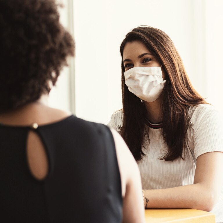 Two individuals are seated at a table, engaged in a conversation. One person is wearing a black top with a unique back design, while the other is dressed in a white shirt with a subtle striped pattern.