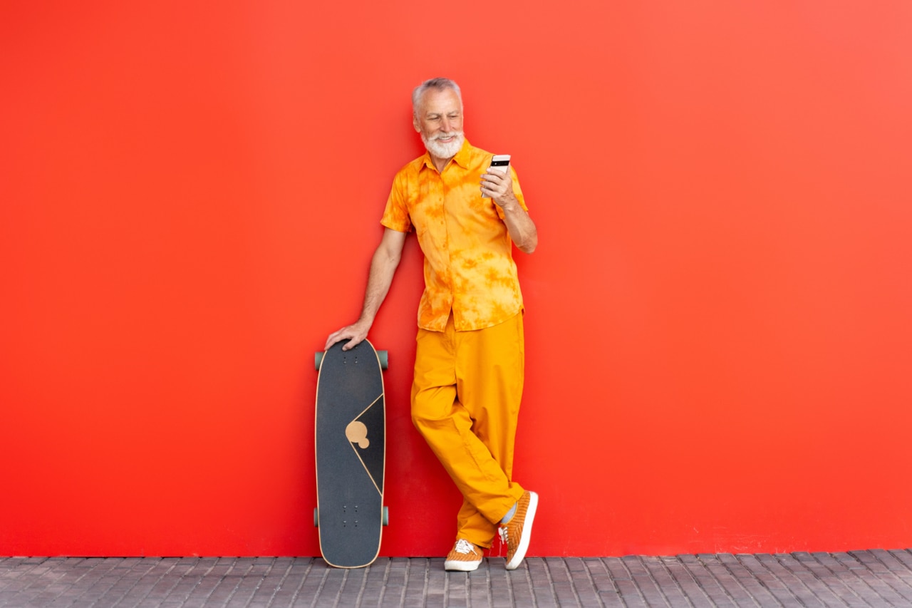 Smiling handsome senior man holding skateboard and mobile phone reading text message, shopping online isolated on red background. Bearded hipster using smartphone, communication online  on the street