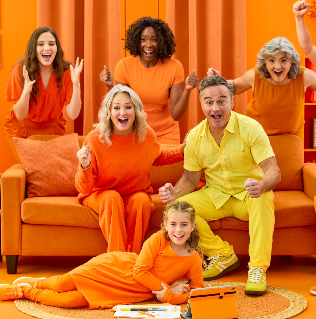 Cheerful group of adults and children in orange and yellow outfits celebrate in a bright orange living room; some sit on an orange sofa, others stand with raised fists, and a child on the floor has a laptop and tablet nearby.
