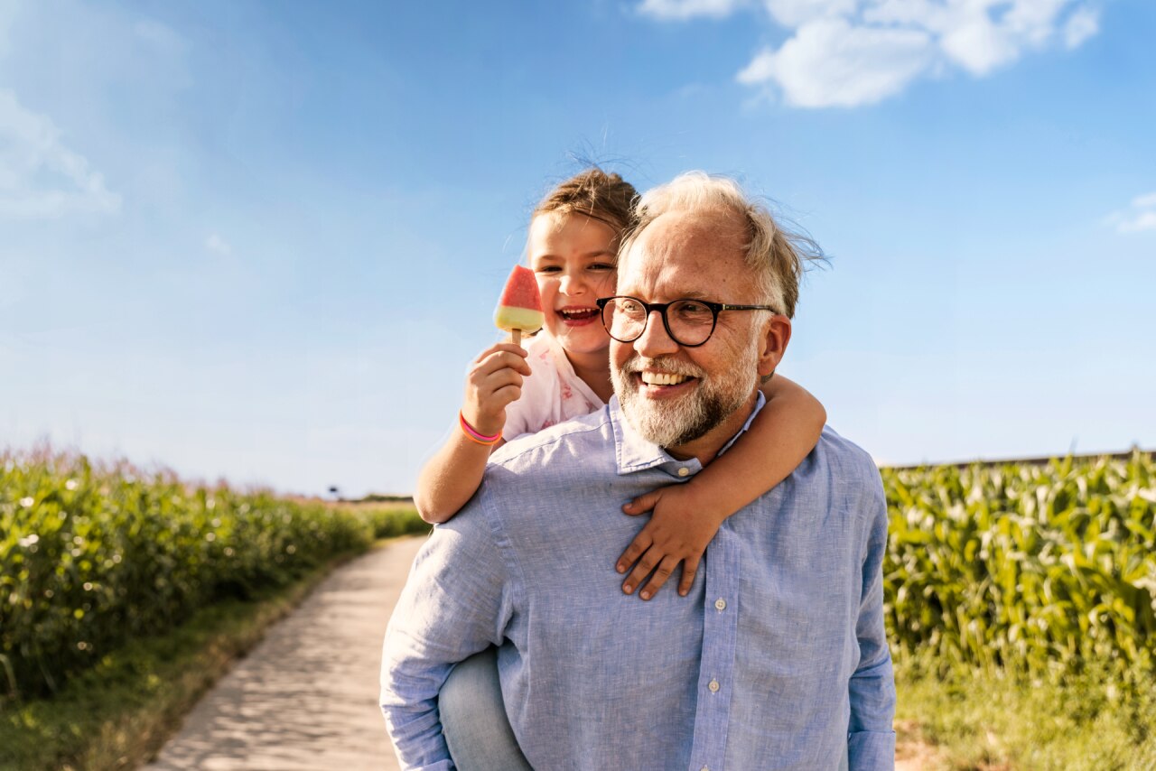 Nonno sorridente che porta la nipotina sulle spalle lungo un sentiero soleggiato attraverso un campo di grano, mentre lei tiene in mano un ghiacciolo all'anguria