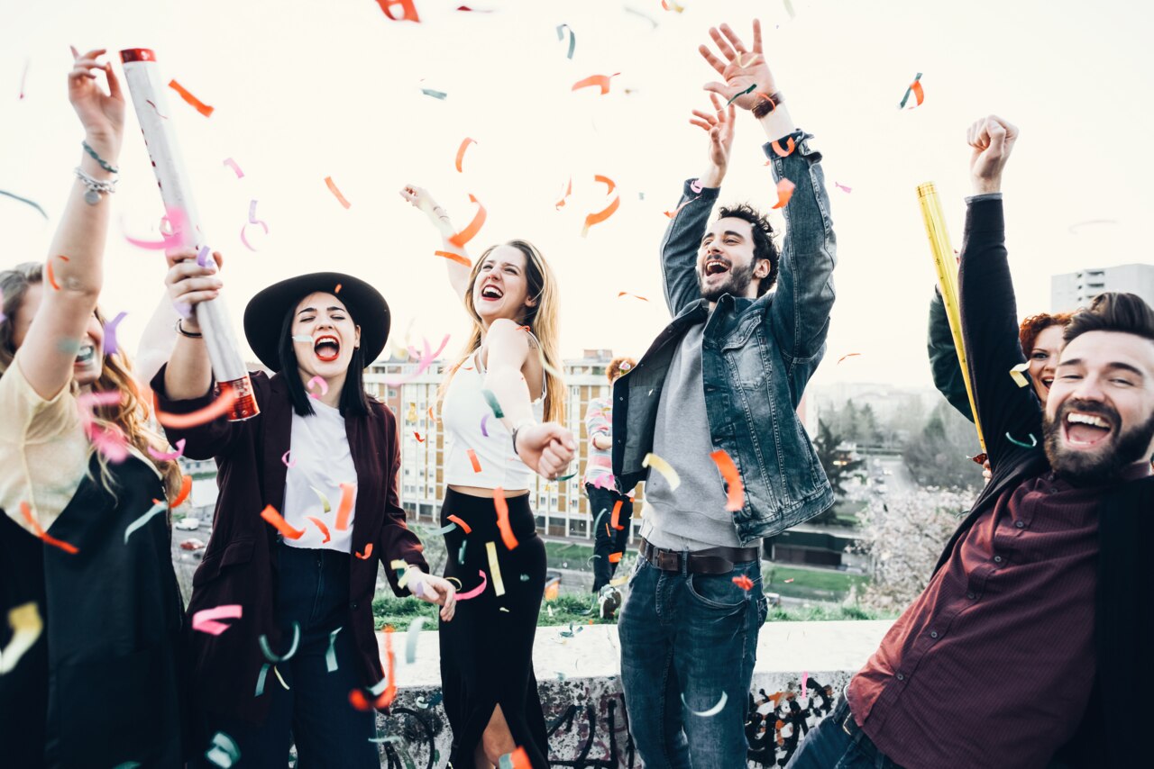 A group of five people is celebrating outdoors, joyfully throwing colorful confetti into the air. They are dressed in casual clothing and are surrounded by a vibrant urban backdrop. The atmosphere is festive and lively, capturing a moment of happiness and celebration.