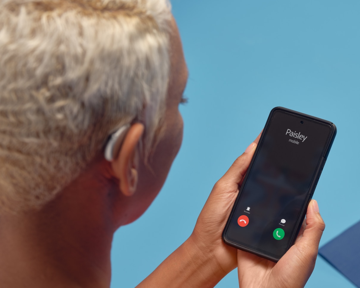 Een Afrikaanse vrouw met wit haar die een hoortoestel draagt en een smartphone vasthoudt waarop ze gebeld wordt, wat erop duidt dat er een verbinding is tussen het hoortoestel en de Remote app.