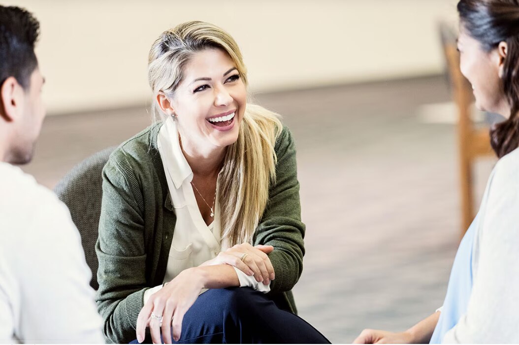 A person is engaged in a conversation with two others in a comfortable indoor setting. The individual is seated, wearing a green cardigan over a white shirt, and appears to be actively listening and participating in the discussion