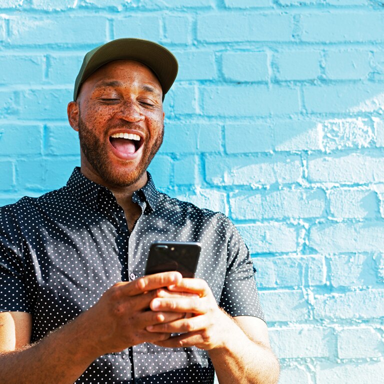 A person wearing a dark polka dot shirt and a green cap is standing in front of a bright blue wall, holding a smartphone in their hands.