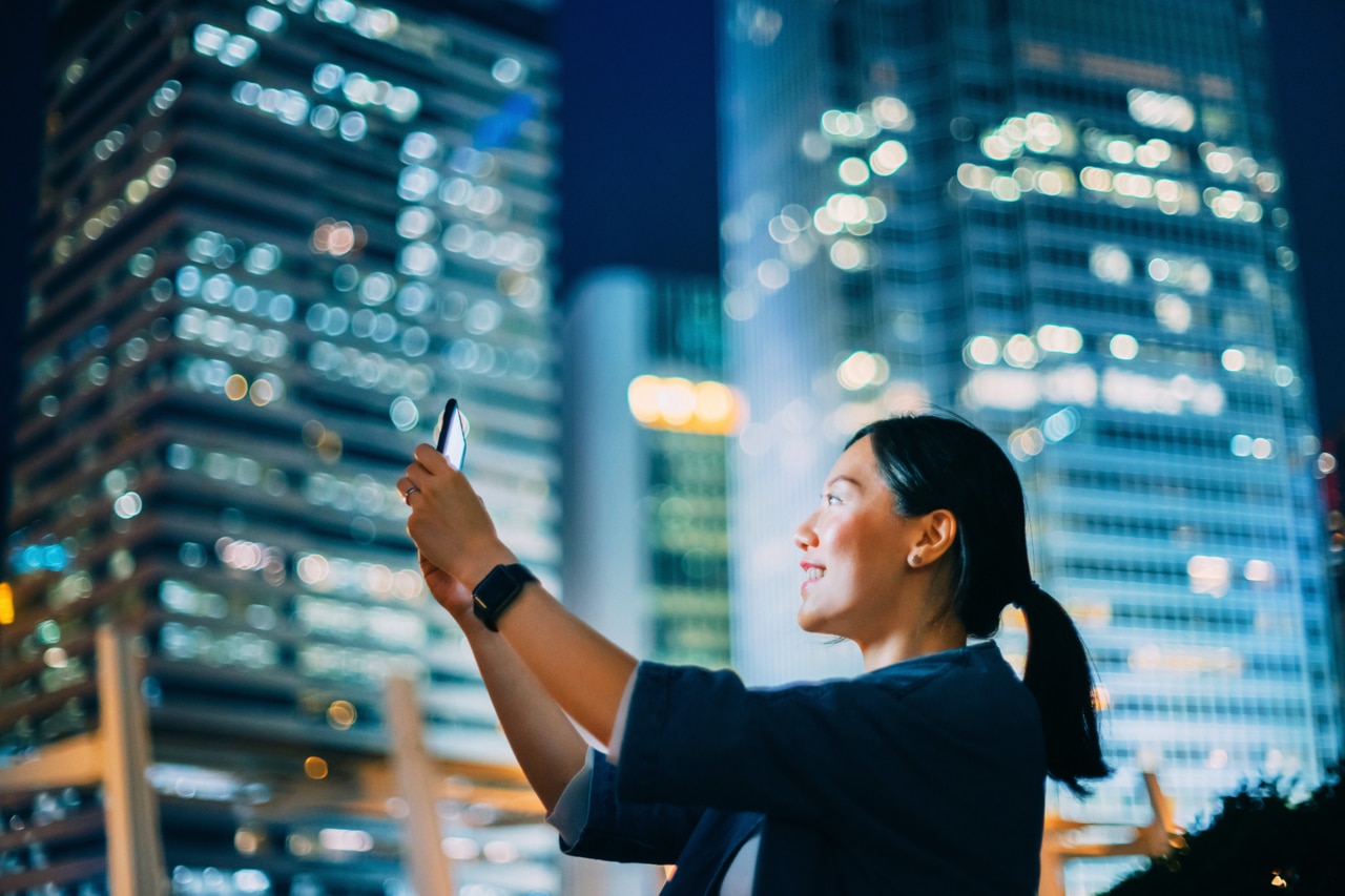 A woman taking a selfie with a smartphone at night, surrounded by a vibrant cityscape. The background features illuminated buildings and city lights, creating a lively urban atmosphere.