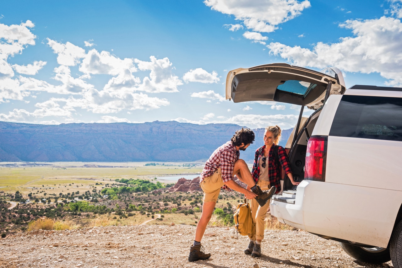 Two young adults preparing for an outdoor adventure by the back of a white SUV parked on a scenic overlook