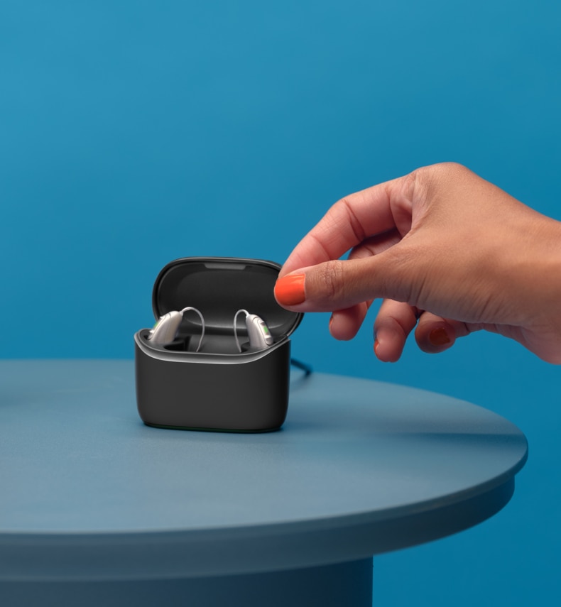 A pair of Vivante hearing aids in a charger standing on a table next to a big red alarm clock
