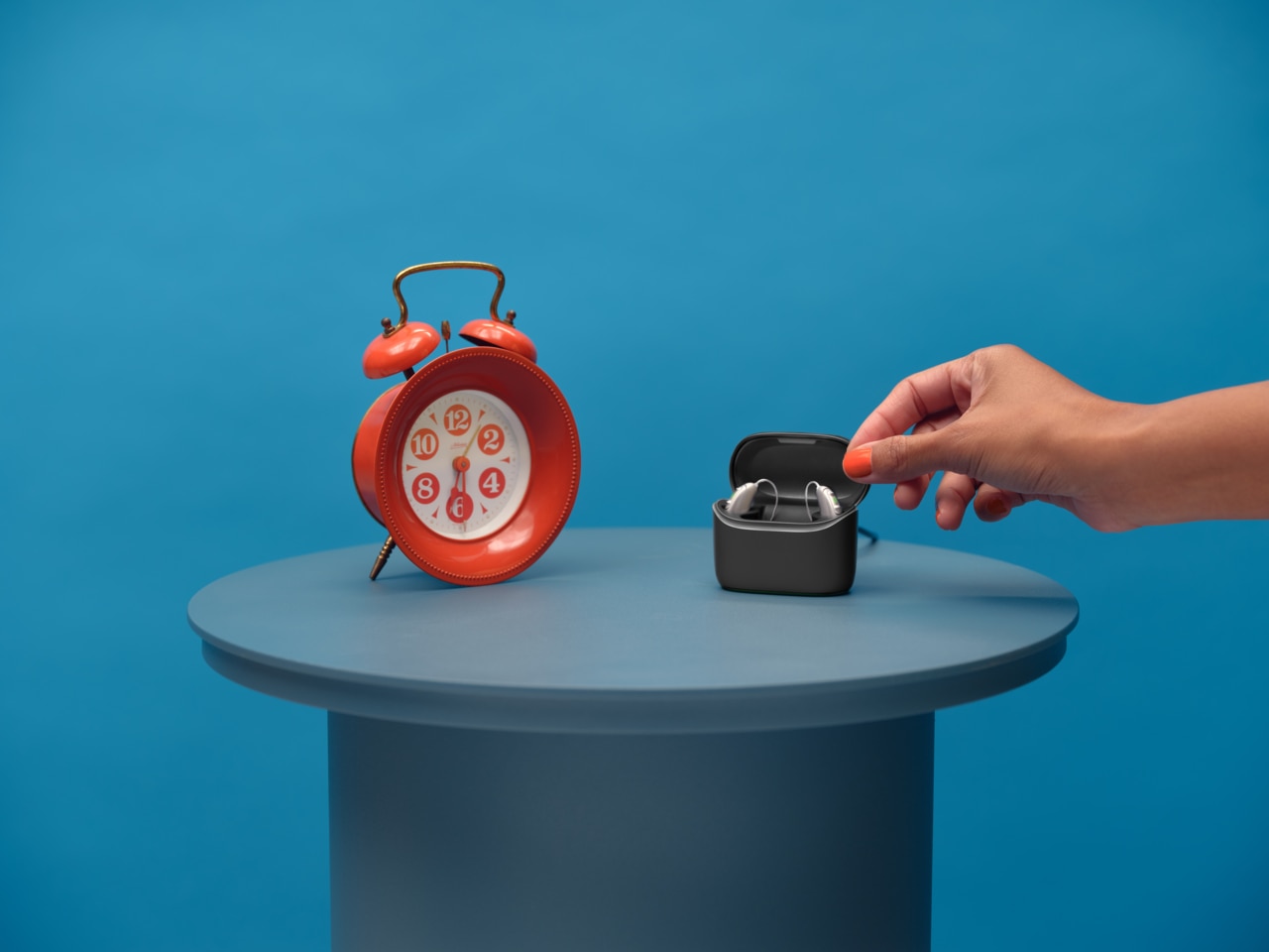 A pair of Vivante hearing aids in a charger standing on a table next to a big red alarm clock