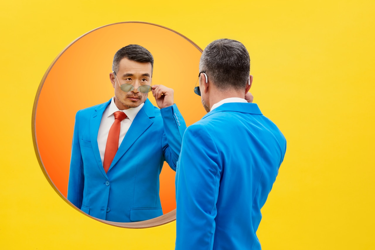 A person in a bright blue suit standing in front of a round mirror against a vibrant yellow background. The individual is adjusting their tie, showcasing a confident and polished appearance. The mirror reflects their stylish outfit, emphasizing the attention to detail in their attire. 