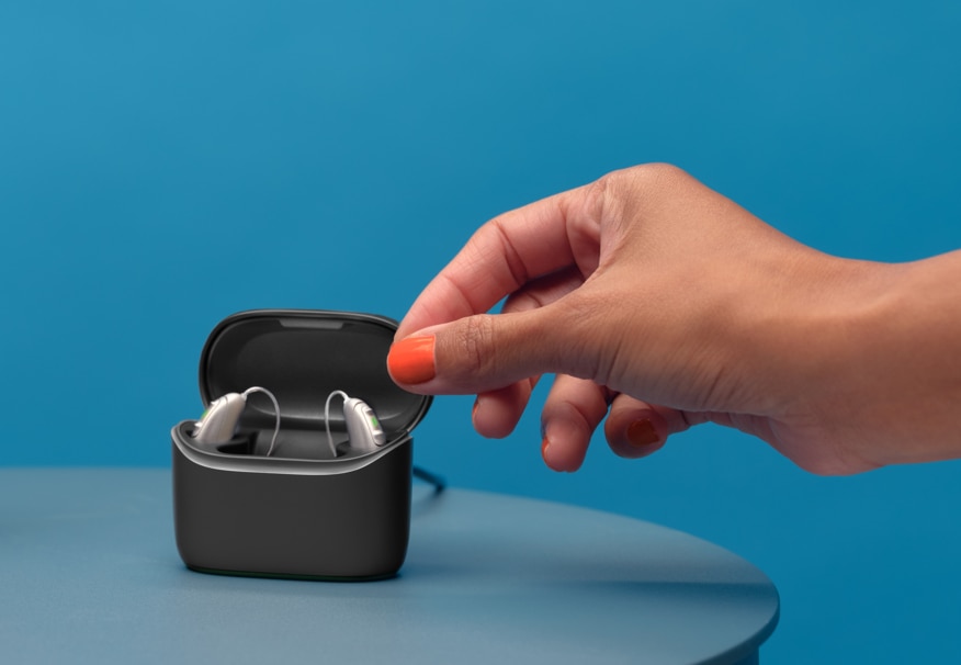 A pair of Vivante hearing aids in a charger standing on a table next to a big red alarm clock