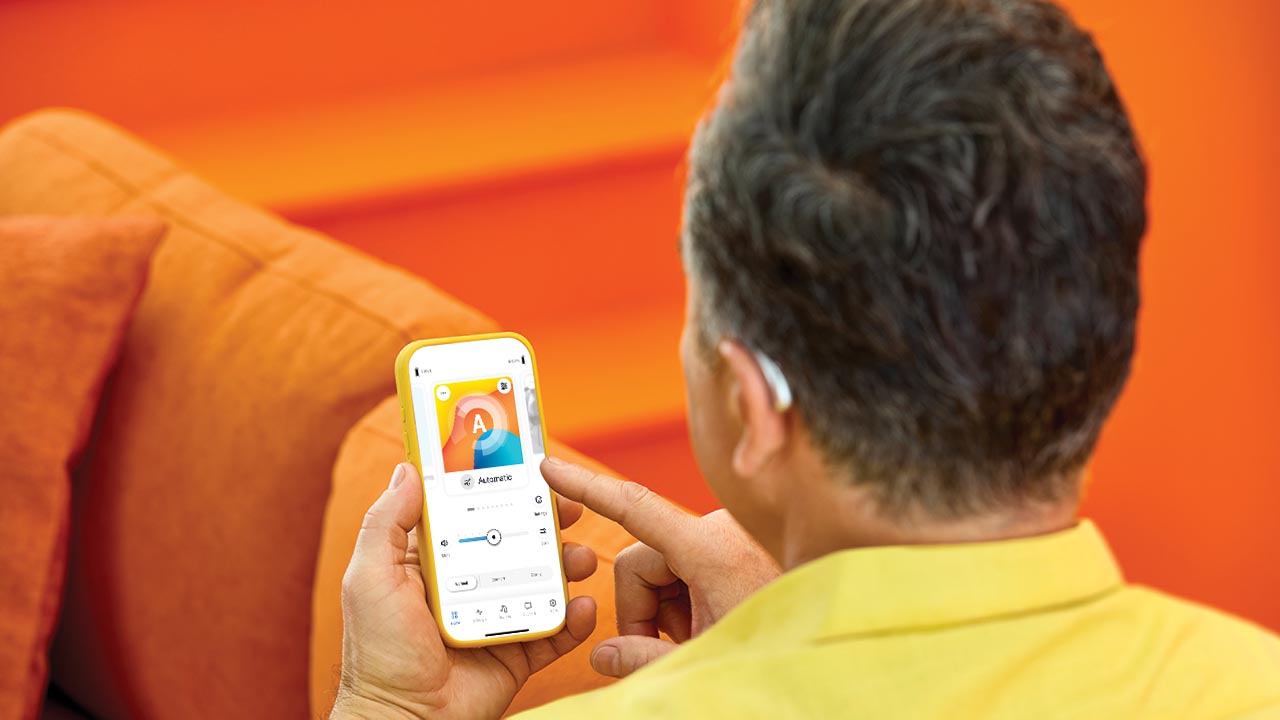Person wearing a Unitron Moxi S-RX hearing aid adjusts settings in the Remote Plus app on a smartphone, showing easy in-the-moment personalization