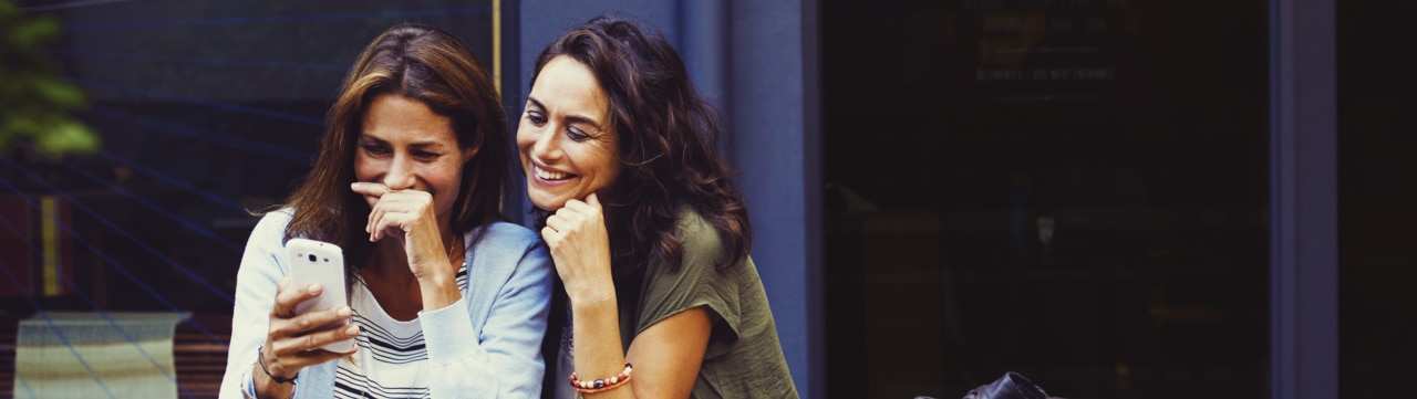 Two women at a cafe laughing while looking at their smartphones