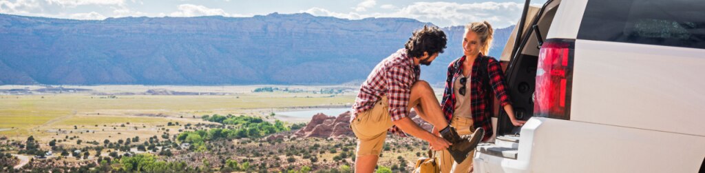Two young adults preparing for an outdoor adventure by the back of a white SUV parked on a scenic overlook